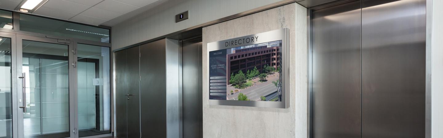 Office building elevator bank with commercial digital signage screen that reads "Directory," showing office photo.